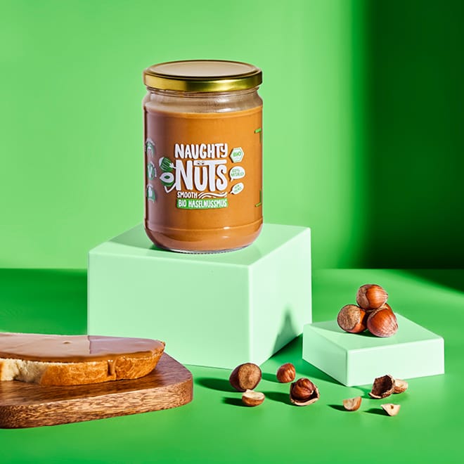 Bio Haselnussmus Smooth (500g Glas) auf einem grünen Podest, mit Haselnüssen und Brot