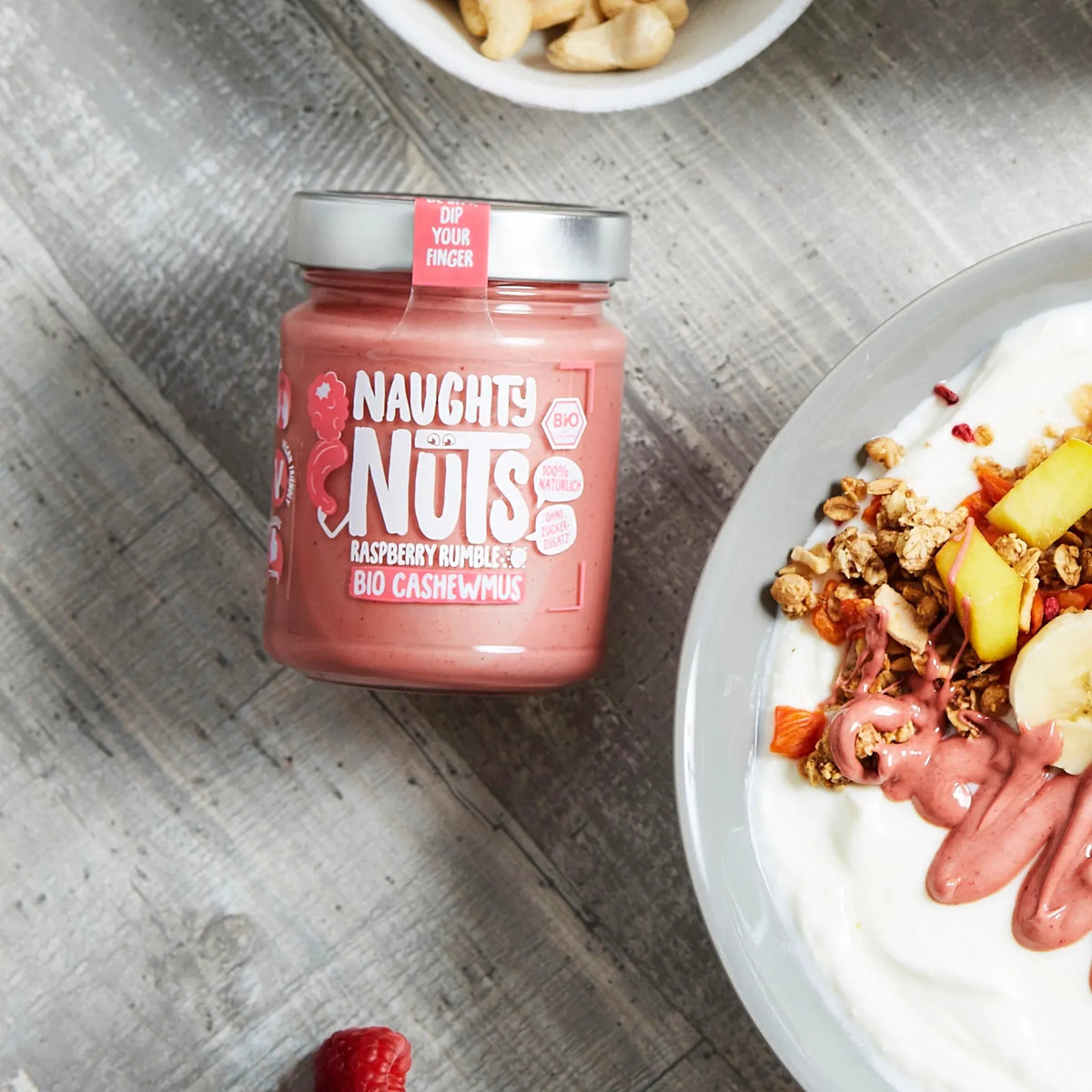 Naughty Nuts Himbeer-Cashewmus neben einer Schale Joghurt mit Müsli und Früchten