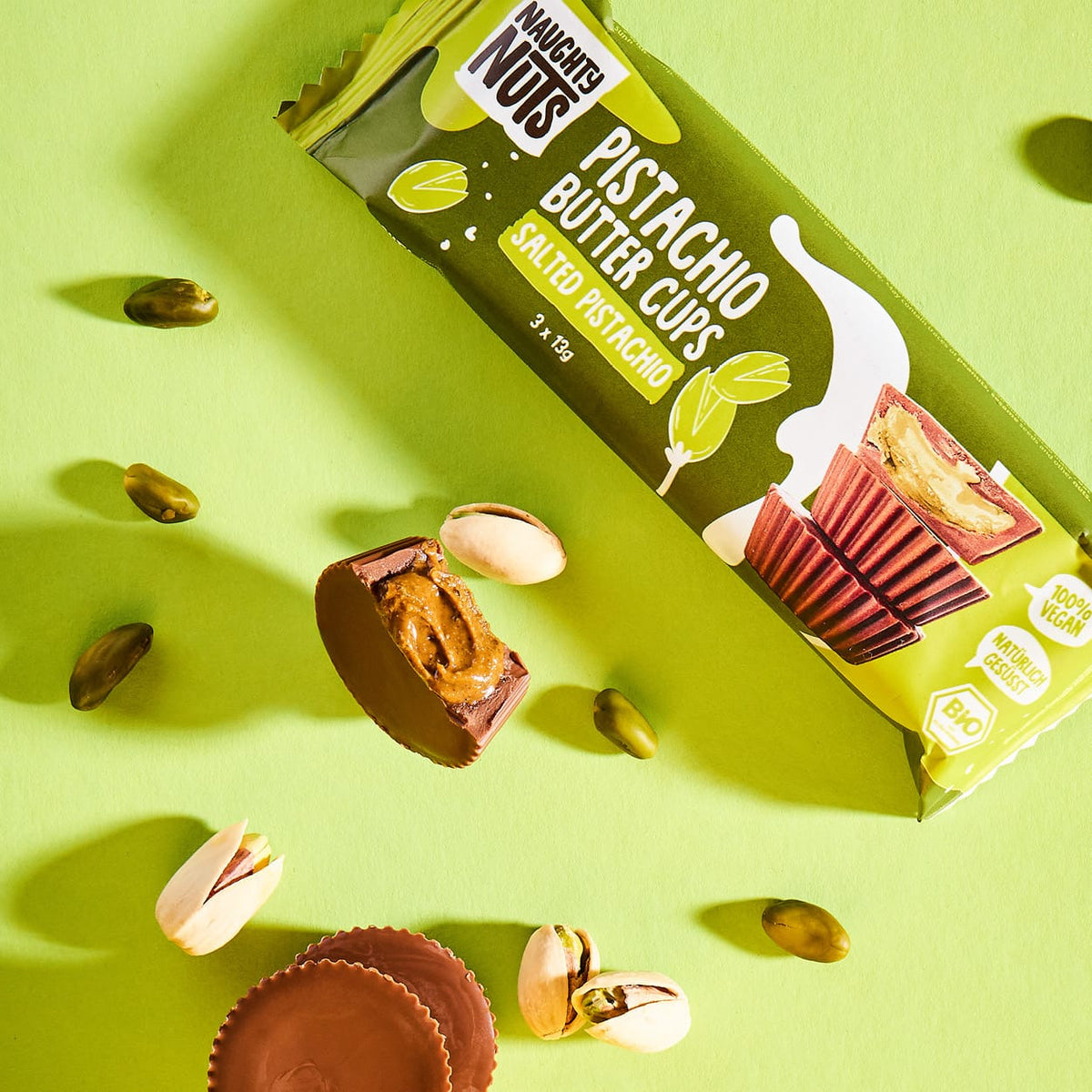 Bio Pistazienbutter Cups Salted Pistachio Packung mit geöffneten Cups und Pistazien auf grünem Hintergrund