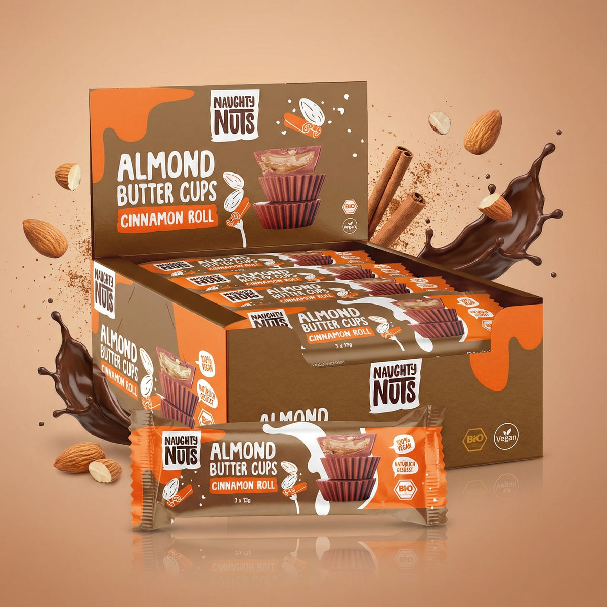 Naughty Nuts Bio Almondbutter Cups Cinnamon Roll Displaybox mit einzelnen Riegeln, Mandeln, Zimtstangen und Schokoladenspritzern