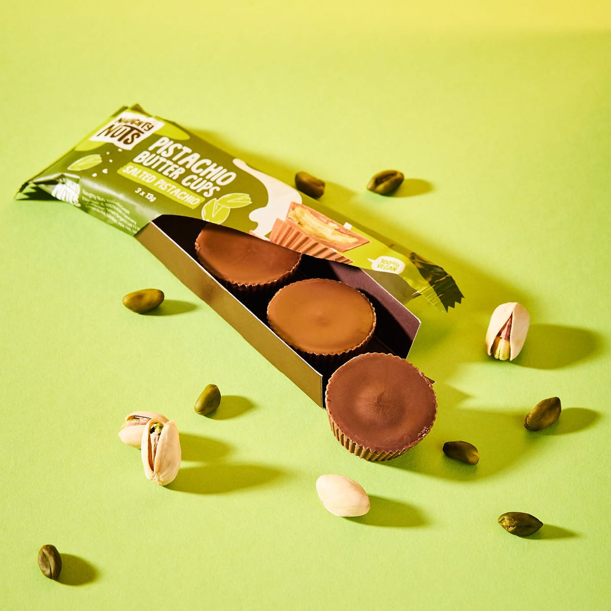 Bio Pistazienbutter Cups Salted Pistachio in offener Verpackung mit einzelnen Pistazien auf grünem Hintergrund