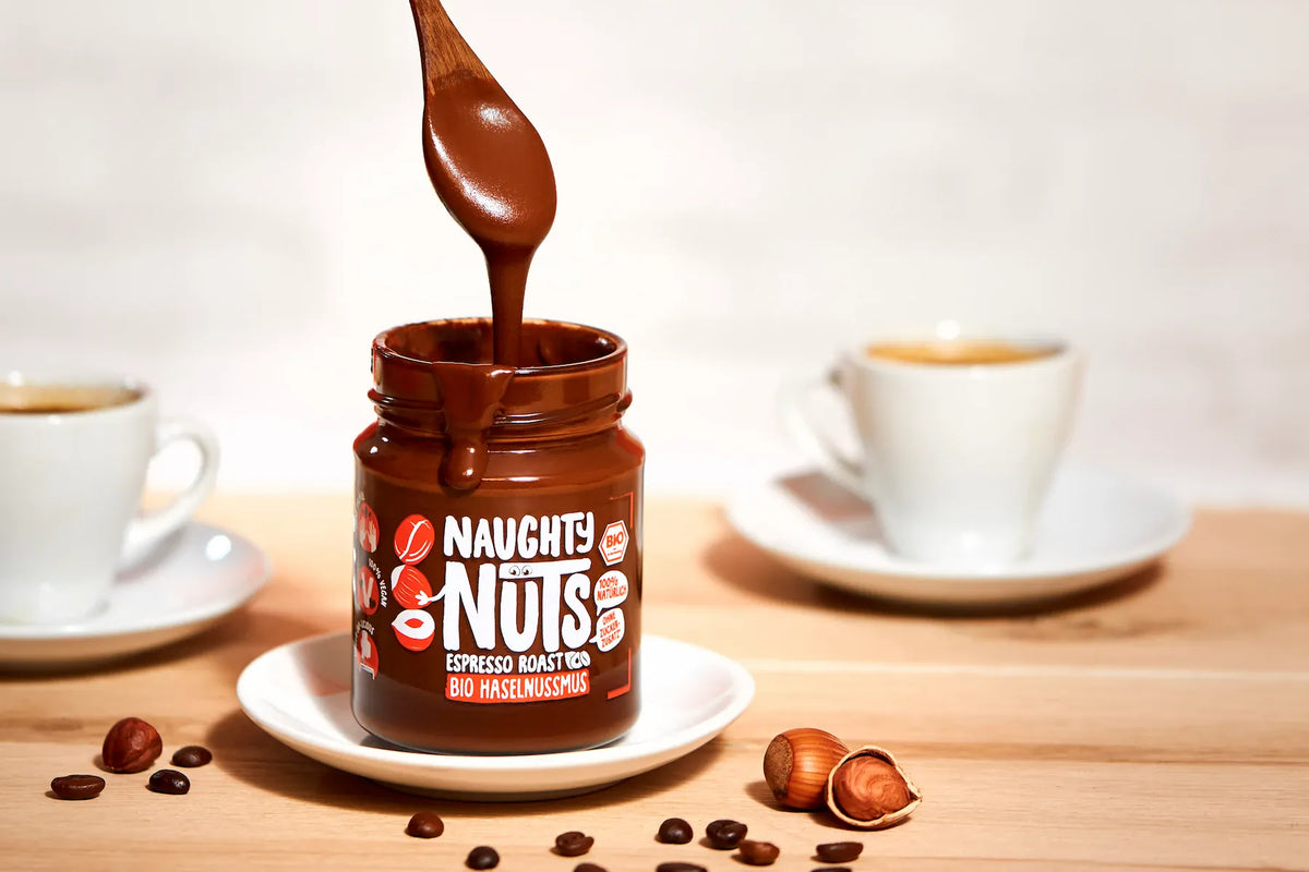 Naughty Nuts Espresso Röstung Bio Haselnussmus im Glas mit Löffel, Kaffeebohnen, Haselnüssen und zwei Tassen Kaffee