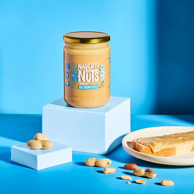 Bio Erdnussmus Crunchy (500g Glas) auf blauem Podest mit Erdnüssen und Toast