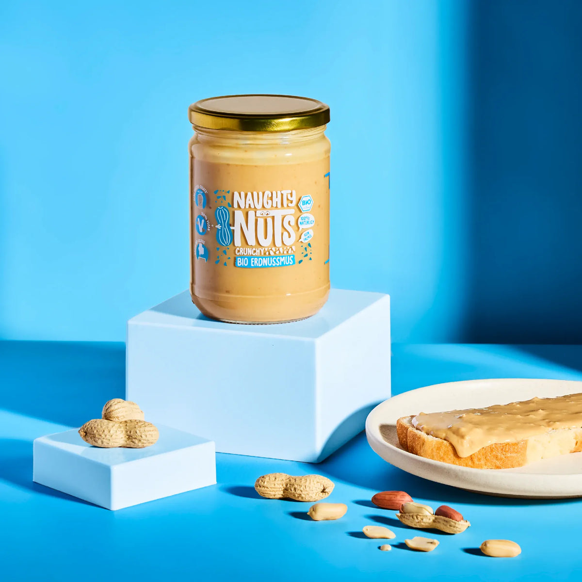 Bio Erdnussmus Crunchy im Glas auf einem Podest mit Erdnüssen und einer Scheibe Brot
