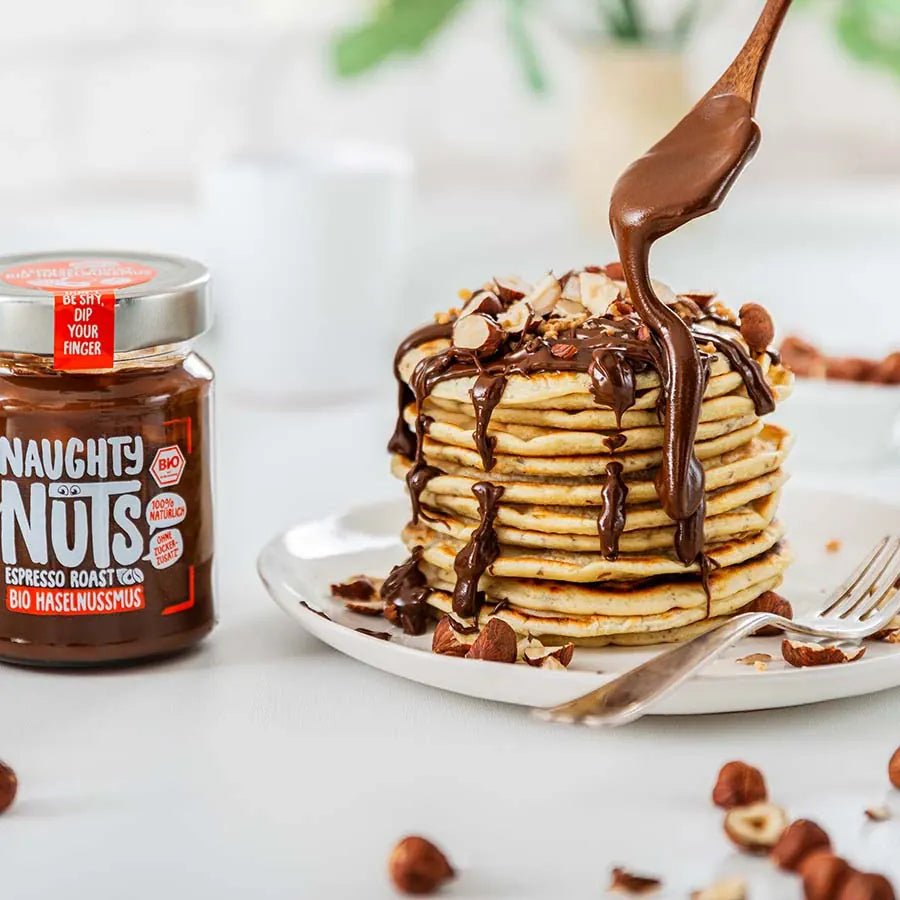 Naughty Nuts Espresso Roast Bio Haselnussmus Glas neben einem Stapel Pfannkuchen mit Schokolade und Haselnüssen