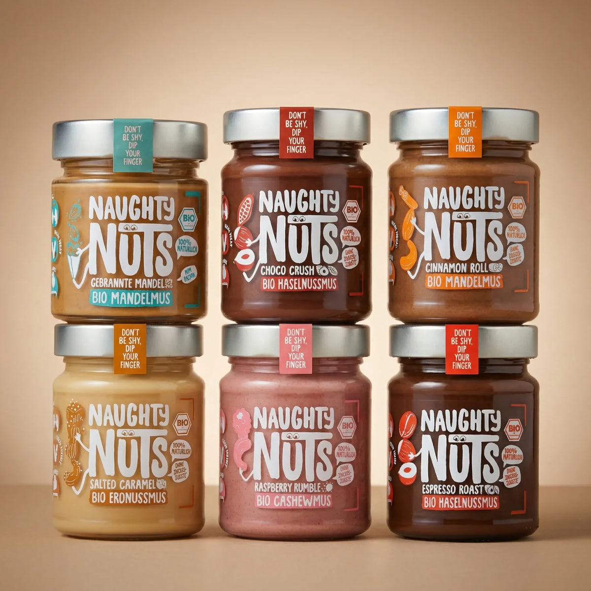 Sechs Gläser Naughty Nuts Nussmus in verschiedenen Geschmacksrichtungen, gestapelt in zwei Reihen