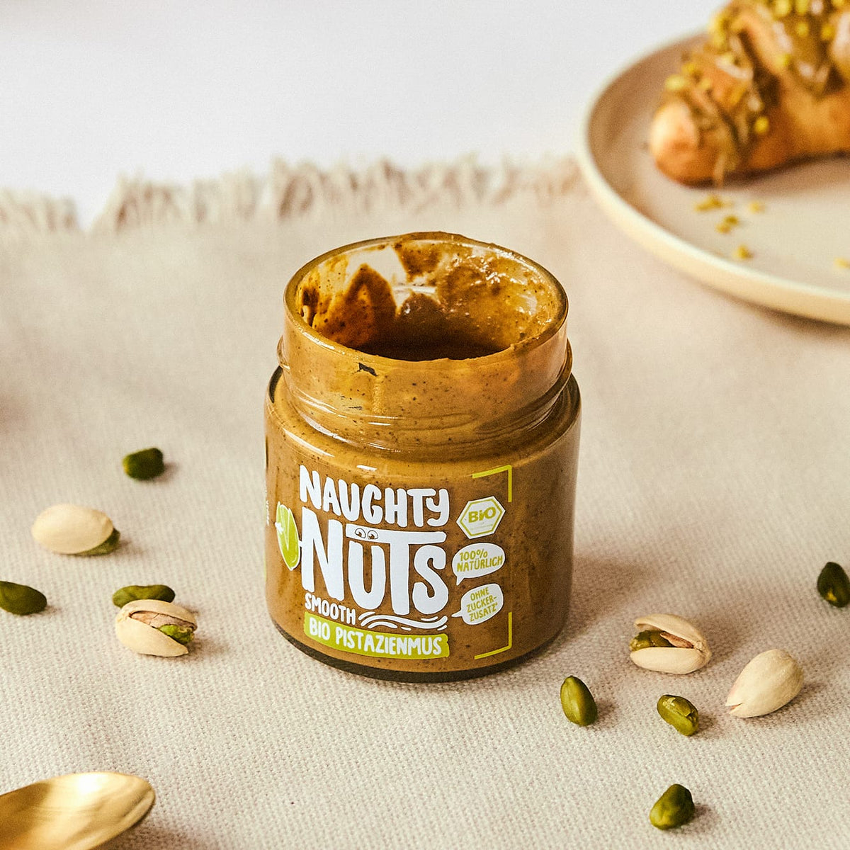 Naughty Nuts Smooth Bio Pistazienmus im Glas mit verstreuten Pistazien und einem Croissant im Hintergrund