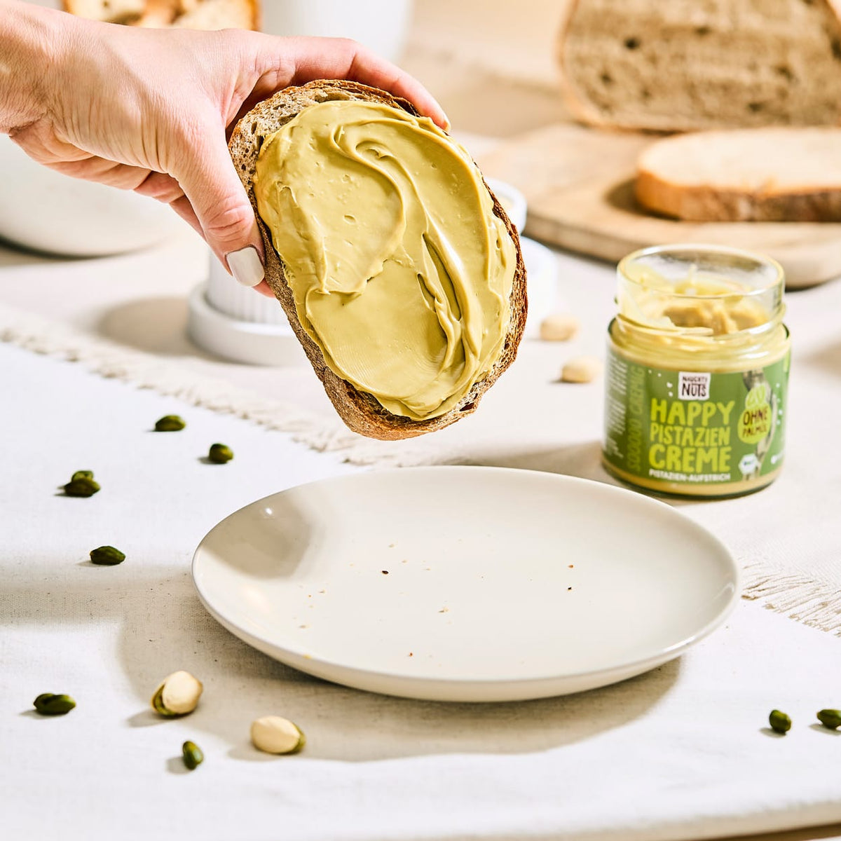 Hand hält ein Brot mit Pistaziencreme über einem Teller, daneben ein Glas Happy Pistazien Creme