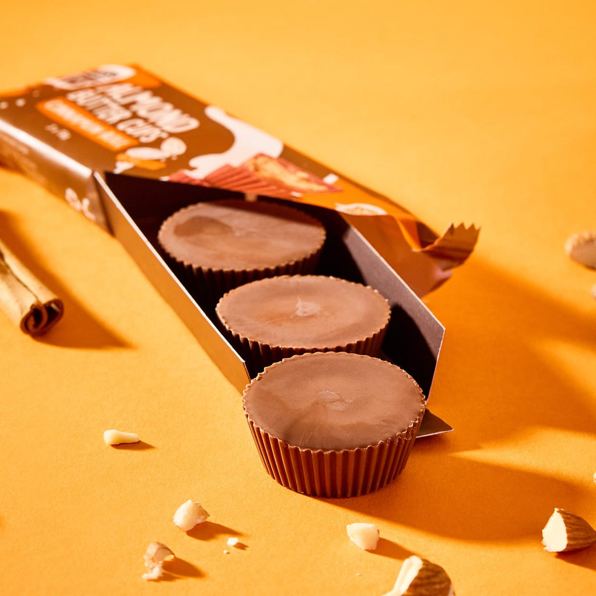 Drei Mandelbutter-Cups in einer offenen Packung auf orangefarbenem Hintergrund mit Mandelsplittern