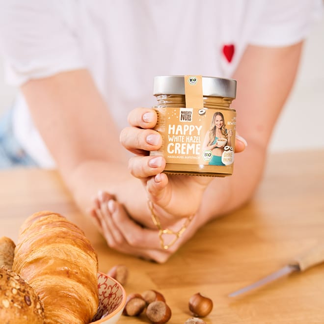 Hand hält ein Glas Happy White Hazel Creme über einem Tisch mit Croissants und Haselnüssen